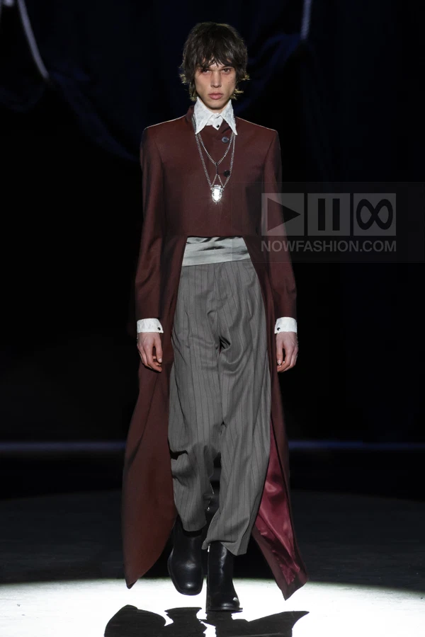Model wearing a long burgundy coat over a white shirt and gray pinstriped trousers on a dark runway.