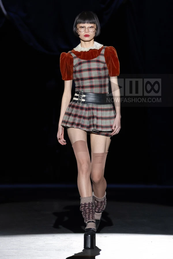 Model on runway wearing a plaid mini dress with velvet sleeves, a wide black leather belt, and platform shoes.