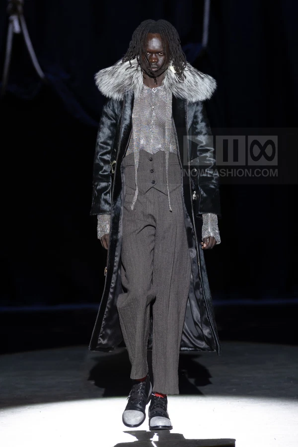 Model walking in a long black fur-trimmed coat, gray waistcoat, and tailored trousers on a dark runway.