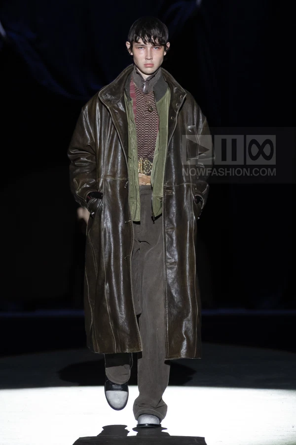 A person walks on a runway wearing a long brown leather coat over a patterned shirt, green vest, and brown trousers.