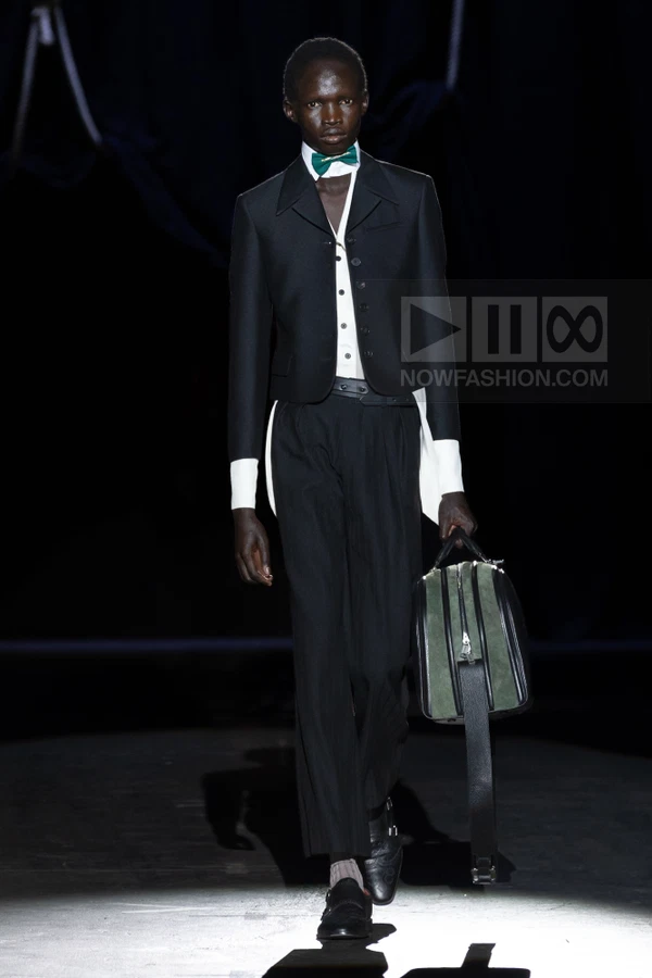 Model wearing a black cropped jacket, white shirt with green bow tie, and black trousers on a dark runway.