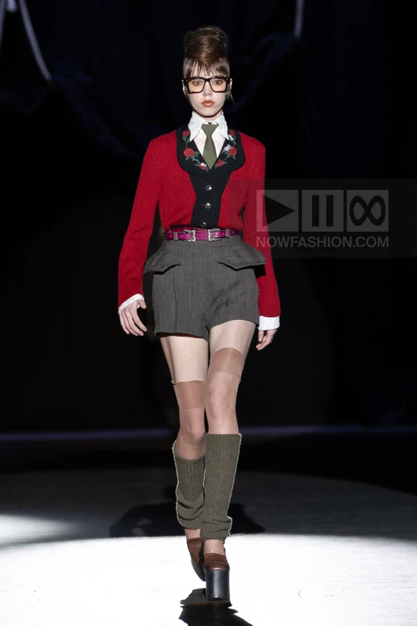 Model on runway wearing a red cardigan with floral embroidery, white shirt, olive tie, gray shorts, and leg warmers.
