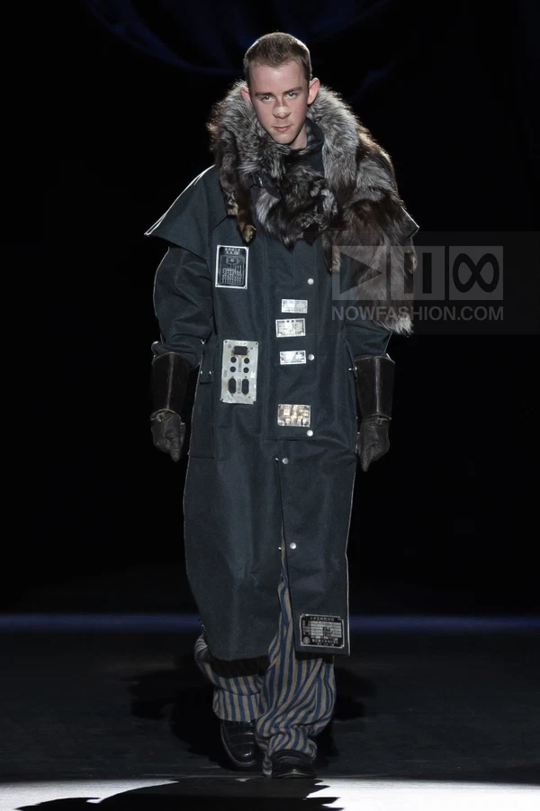 Model wearing a long dark coat with fur collar, metal patches, and striped trousers on a dark runway.