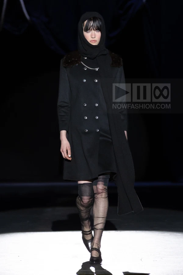 A person walks in a black double-breasted coat, sheer tights, and a headscarf on a dark runway.