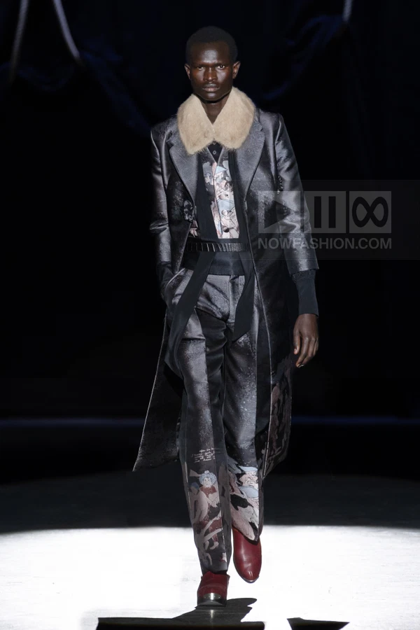 A model walks the runway wearing a dark patterned coat with a fur collar, matching trousers, and burgundy boots.