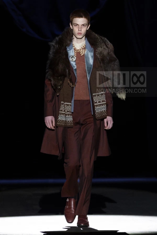 A person walks down a runway wearing a dark brown coat, fur stole, brown sweater, blue shirt, and brown trousers.
