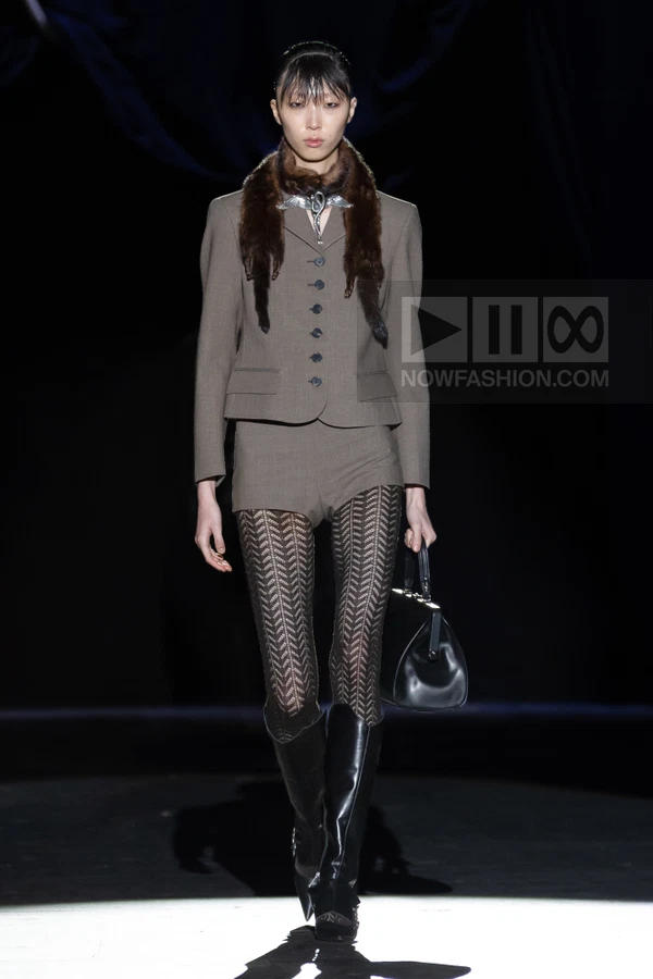 Model wearing a brown tailored jacket with fur collar, matching shorts, patterned tights, and black boots on a runway.