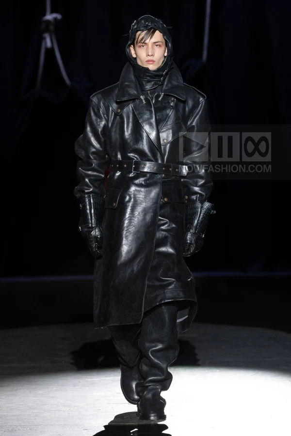 A person wearing a long black leather trench coat, matching leather trousers, and a hood, walking on a runway.