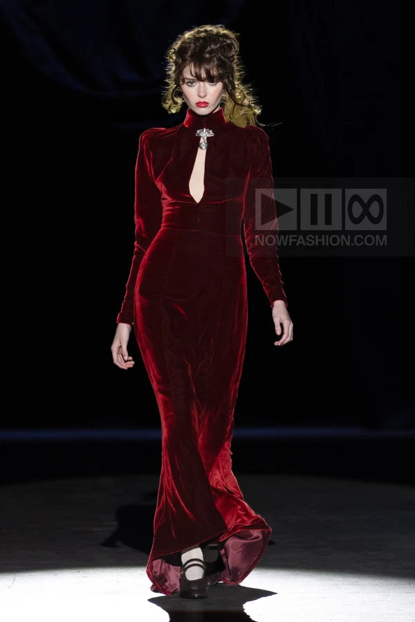 A person walks in a floor-length dark red velvet gown with a keyhole neckline and a silver brooch at the collar.