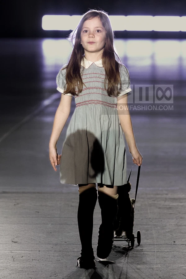A child walks down a runway wearing a gray smocked dress with a white collar and black knee-high socks.