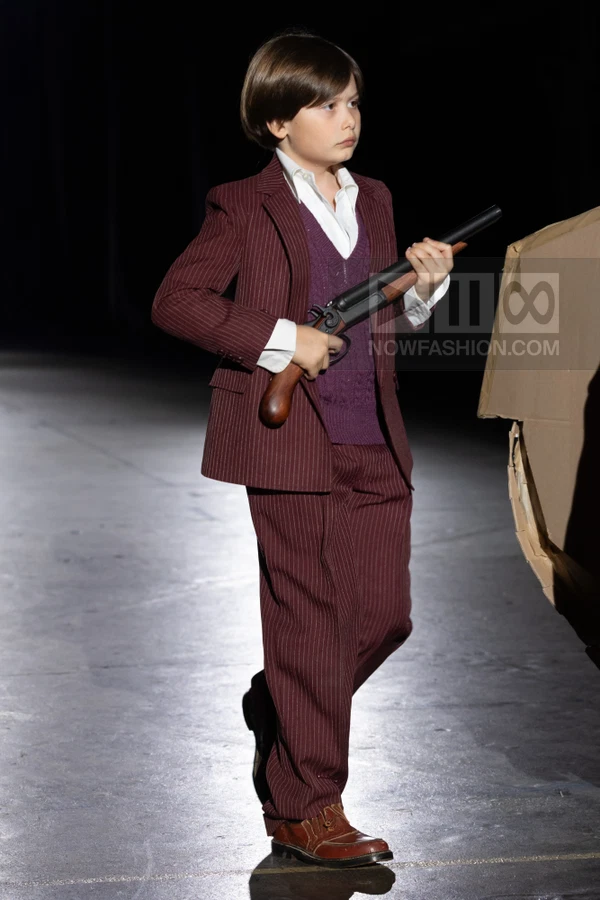 A person wearing a burgundy pinstripe suit, purple sweater vest, and white shirt, holding a prop shotgun on a runway.