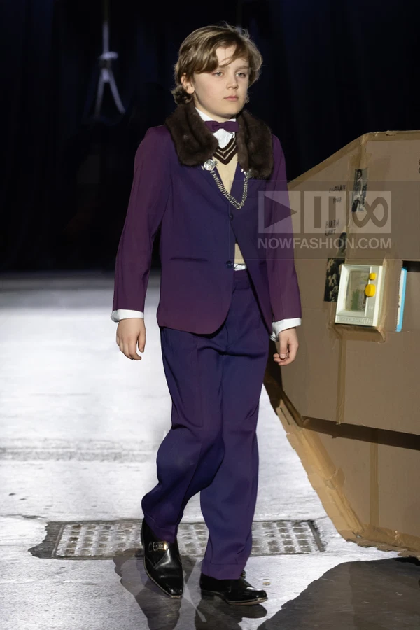 A person wearing a purple suit with a fur collar, a bow tie, and a sweater vest walks on a runway.