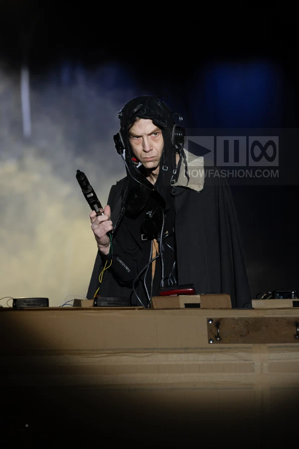 A person wearing a black cape and headgear holds a prop gun behind a cardboard structure on a dark runway.