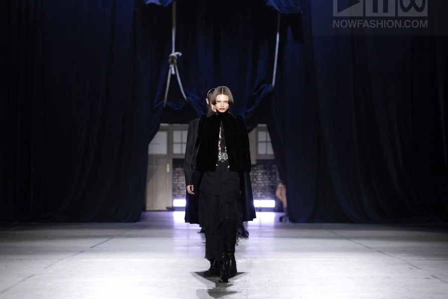 Model walking on runway wearing black velvet jacket and layered skirt with dark background.