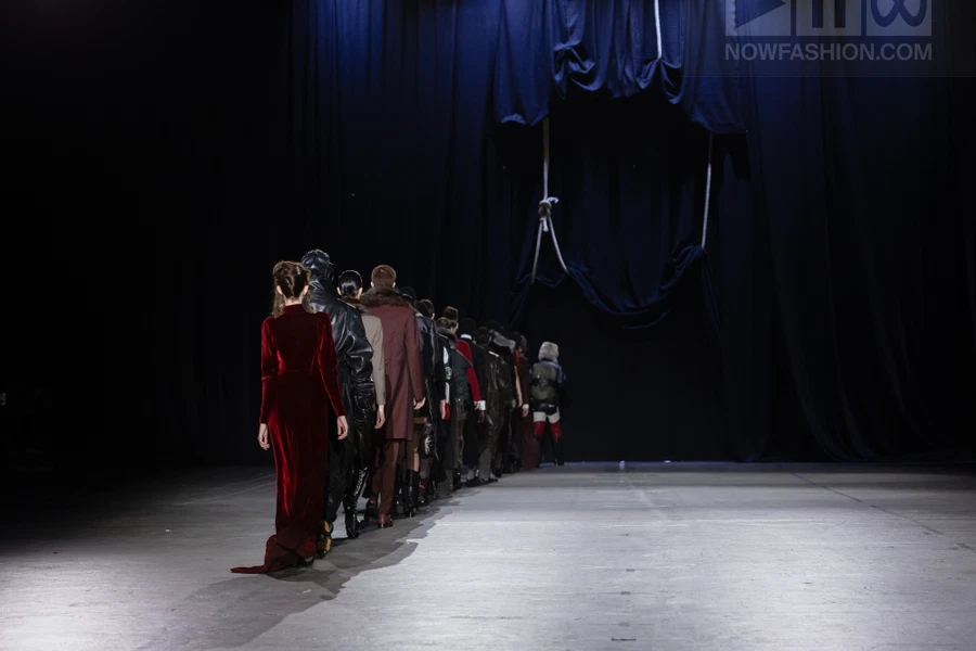 Models walking away in a line on a dark runway towards a draped curtain backdrop.