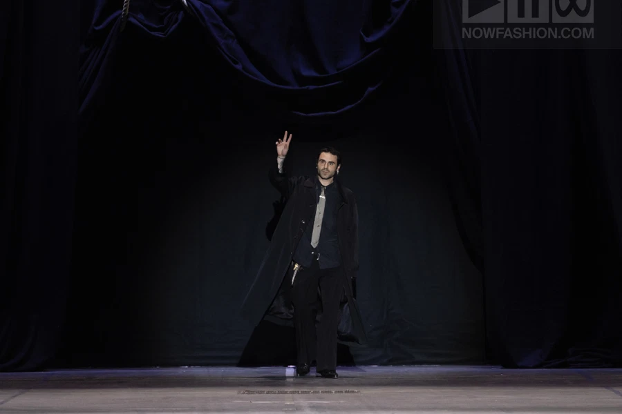 A person in a dark coat and trousers walks on a stage against a dark curtain background.
