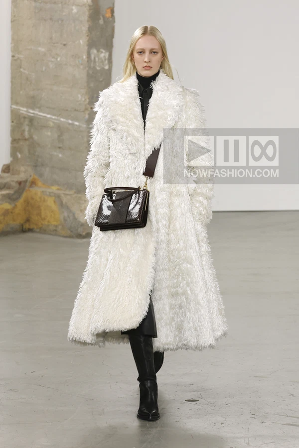 Model wearing a long white shaggy coat over a black outfit with black boots and a dark brown cross-body bag on a runway.