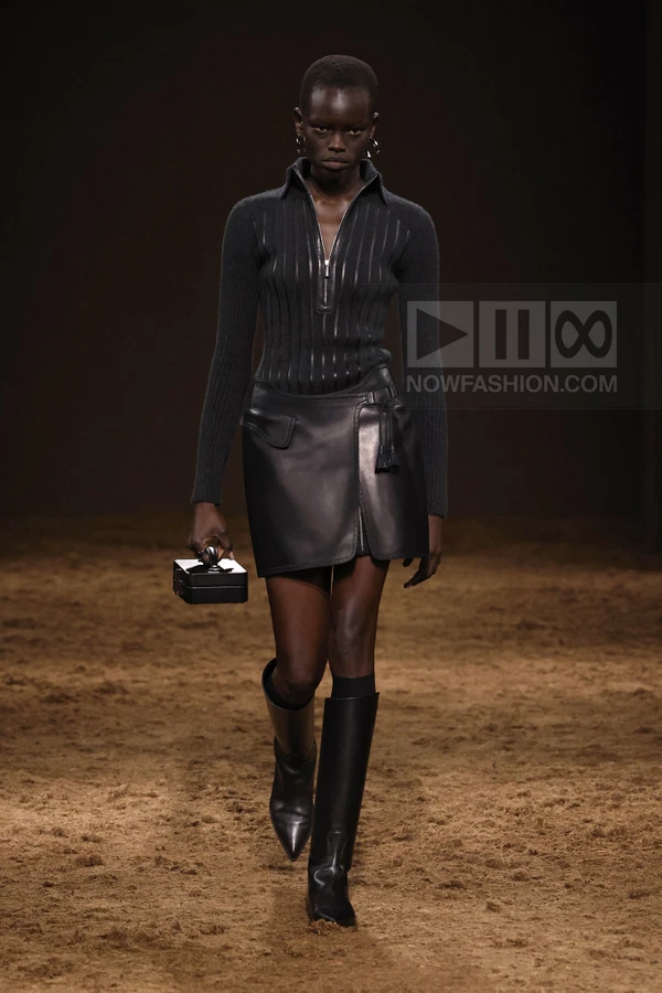 Model walking in a black ribbed half-zip sweater, a leather wrap mini skirt, and knee-high boots, carrying a box bag.