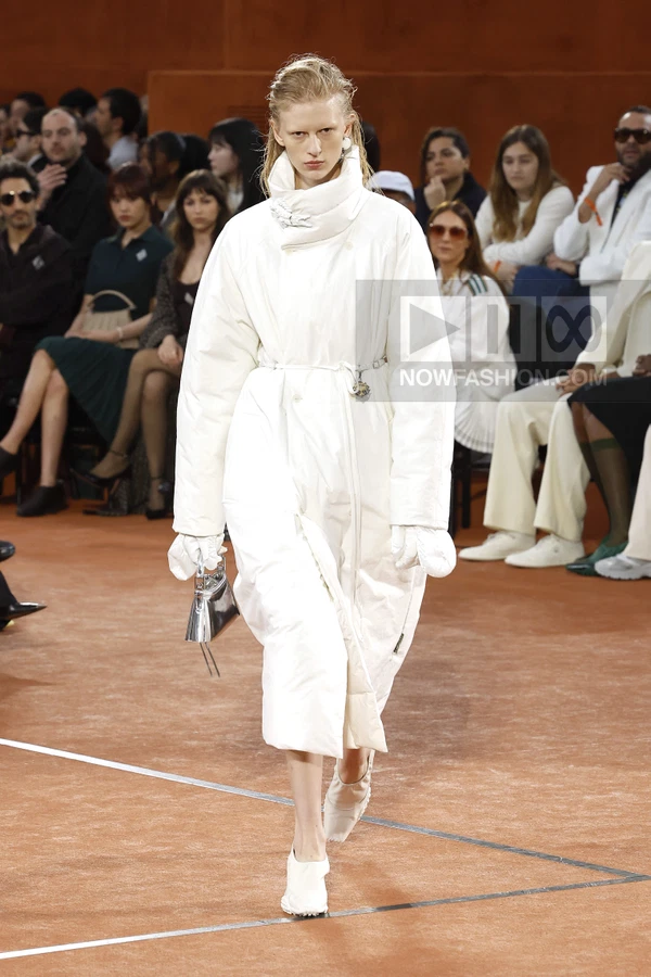 Model walking on a clay-colored runway wearing a white puffer coat with a high collar, white gloves, and white shoes.