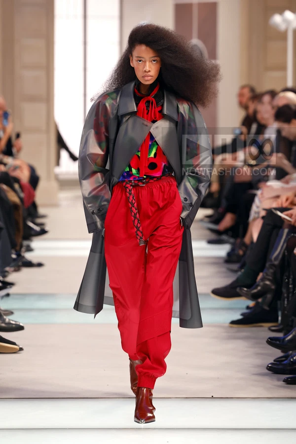 Model on runway wearing a translucent gray trench coat over a red patterned top and bright red trousers with a braided belt.