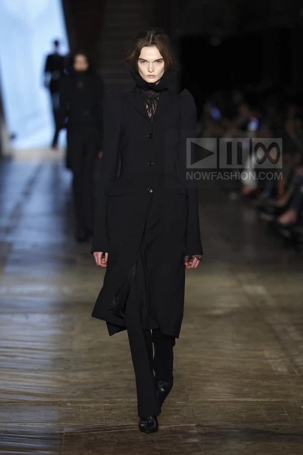 A model walks the runway in a black tailored coat with structured shoulders, worn over black trousers and lace top.