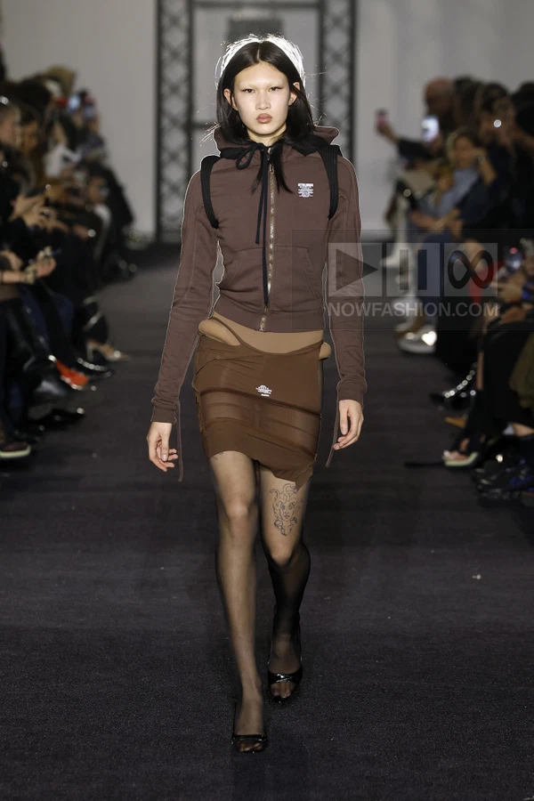 Model walking in a brown zip-up hoodie, layered mini skirt, and sheer tights on a dark runway.