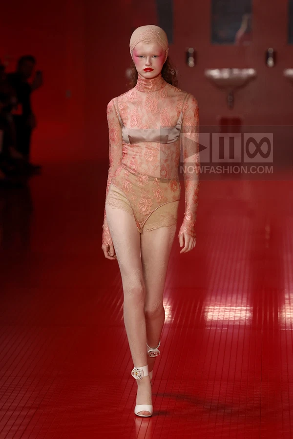Model wearing a sheer lace bodysuit with a satin bra overlay and matching headcap on a red runway.