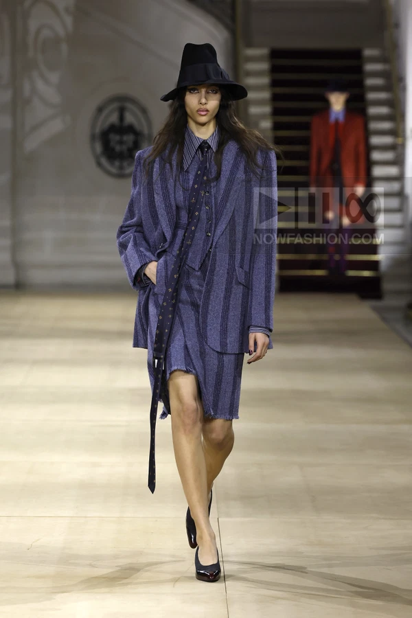 Model walking in a purple striped wool blazer and skirt suit with a black hat, tie, and dark pumps on a runway.