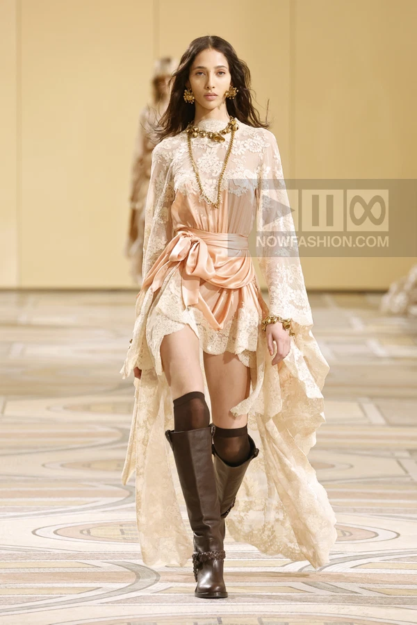 Model walking in a cream lace dress with a satin waist tie, gold chain necklace, and dark brown knee-high boots.