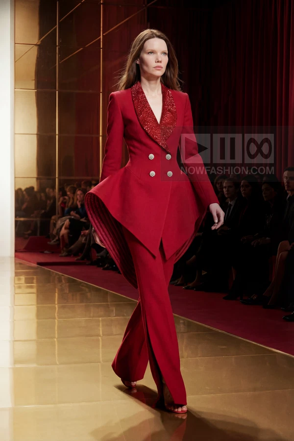 A model walks the runway in a red double-breasted jacket with a peplum hem and matching red trousers.