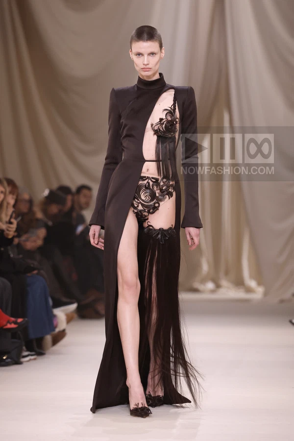 Model on runway wearing black gown with cut-outs, intricate dark embellishments, and long fringe details.