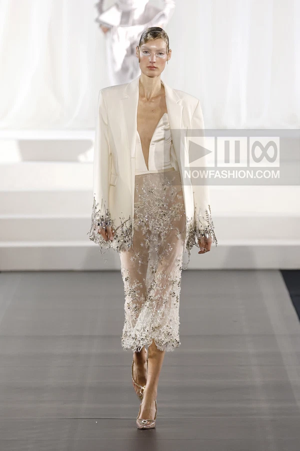 Model wearing a white blazer with crystal fringe over a sheer, beaded midi skirt and white bodice on a runway.