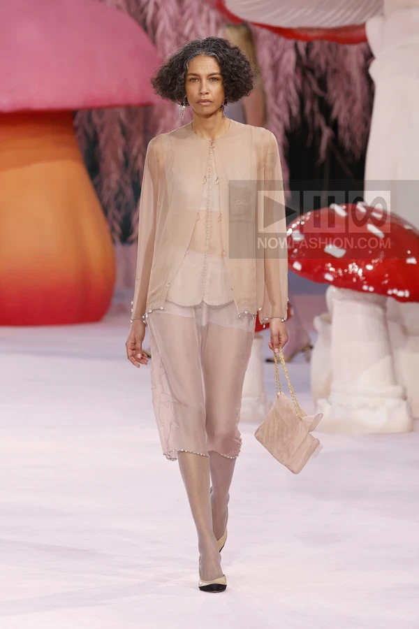 Model walking in a sheer beige jacket and matching midi skirt with a small quilted chain bag on a runway.