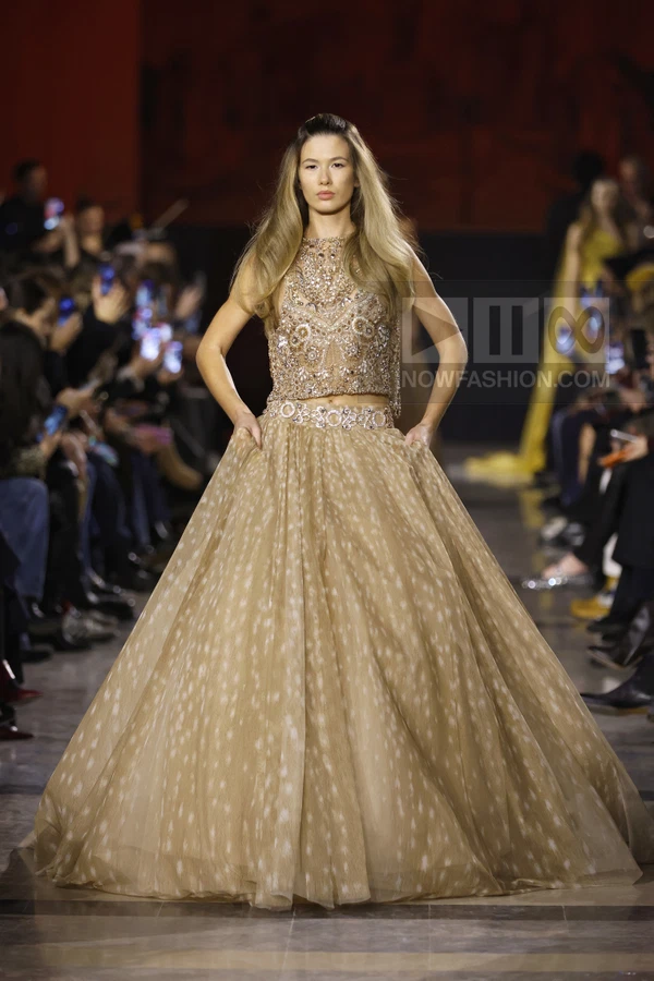 A model on a runway wears a beige two-piece gown with a heavily beaded crop top and a voluminous floor-length skirt.