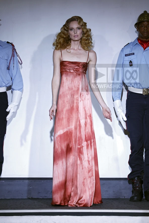 A model in a floor-length red and white tie-dye silk dress stands between two figures in uniform on a runway.