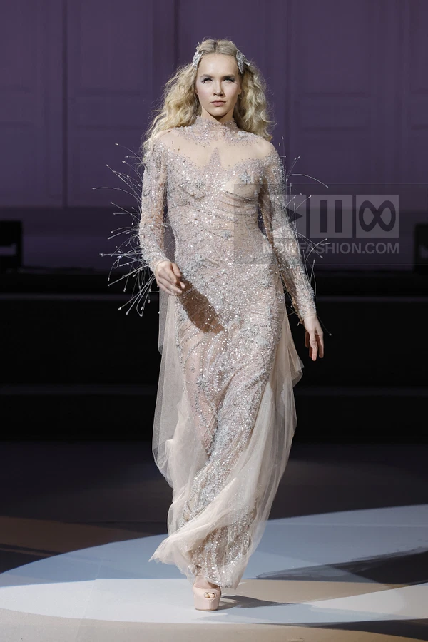 A model walks the runway in a sheer, floor-length gown covered in silver sequins and delicate wire embellishments.