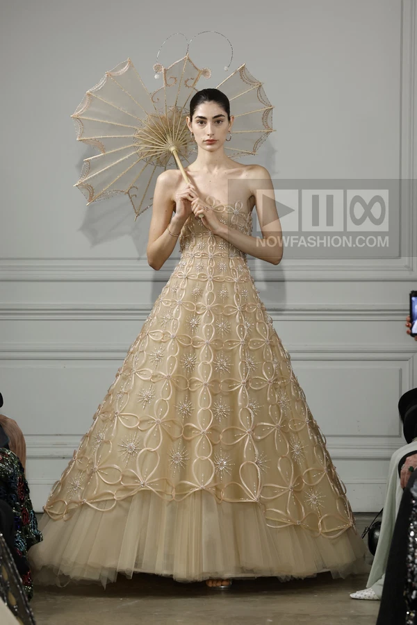 Model in a beige strapless gown with geometric ribbon embroidery and a matching parasol on a runway.
