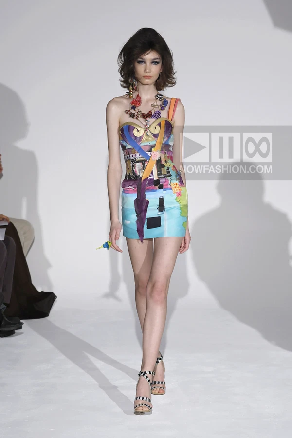 Model wearing a colorful, patchwork-style mini dress with a bustier bodice and graphic prints on a white runway.