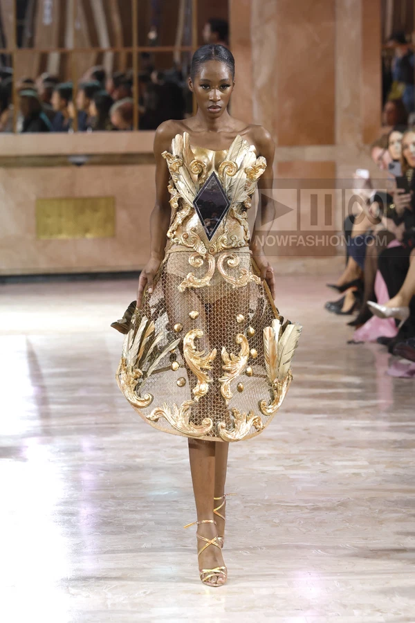 Model wearing a gold metallic structured strapless dress with a wire mesh skirt and ornate scrollwork on a runway.