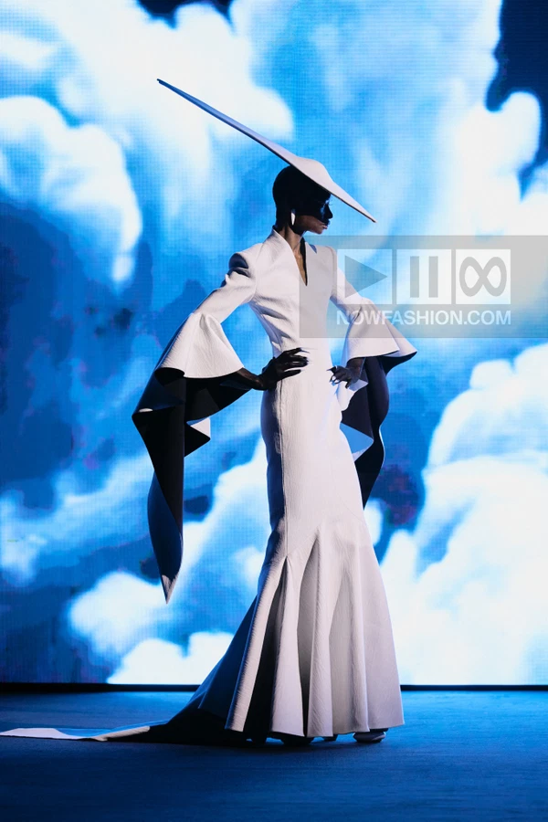 A person in a white gown with flared sleeves and a wide-brimmed hat stands against a blue cloud-patterned backdrop.