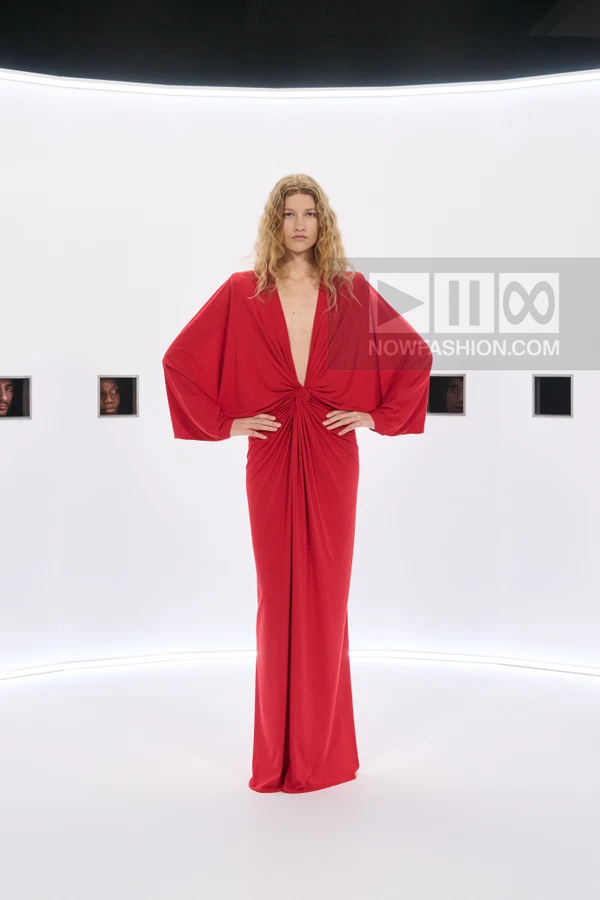 A model wearing a floor-length red gown with a deep V-neckline and wide sleeves stands against a white backdrop.