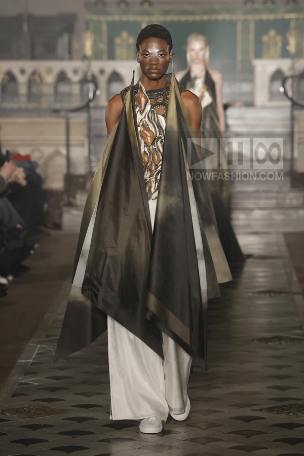 Model walking in a layered, dark-toned, architectural garment with a white skirt and white sneakers on a runway.