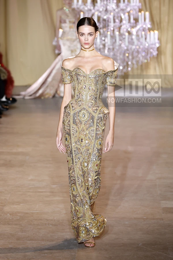 A model walks the runway in a gold, heavily embellished, off-the-shoulder gown with a corset-style bodice.