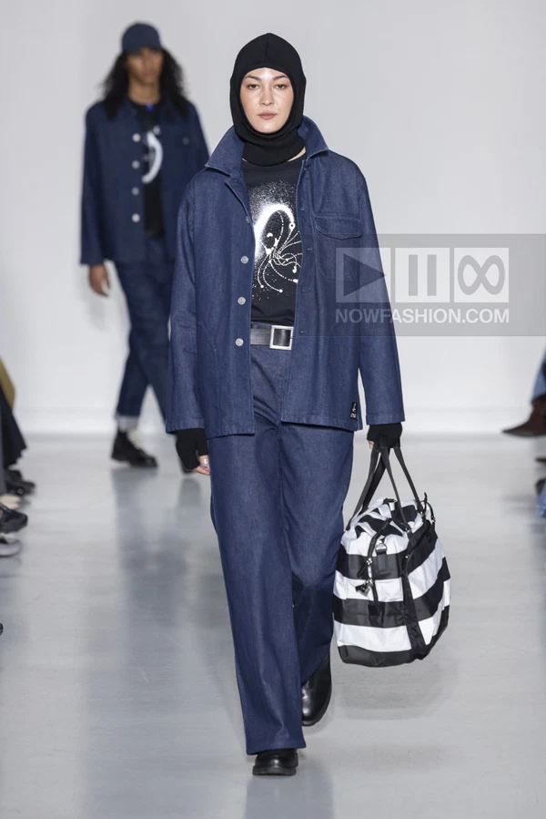 Model walking in dark blue denim jacket and wide-leg trousers, black balaclava, and black and white striped duffel bag.