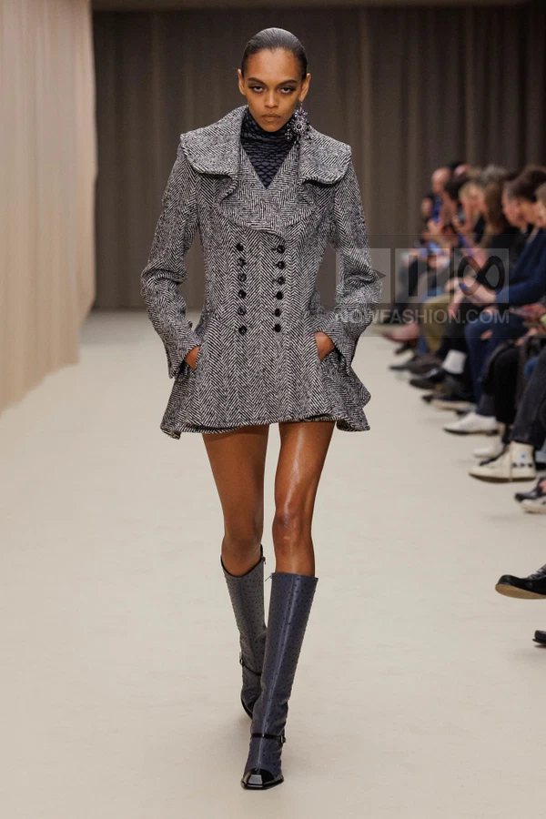 Model walking in a herringbone double-breasted coat with a flared hem, worn over a dark turtleneck and knee-high boots.