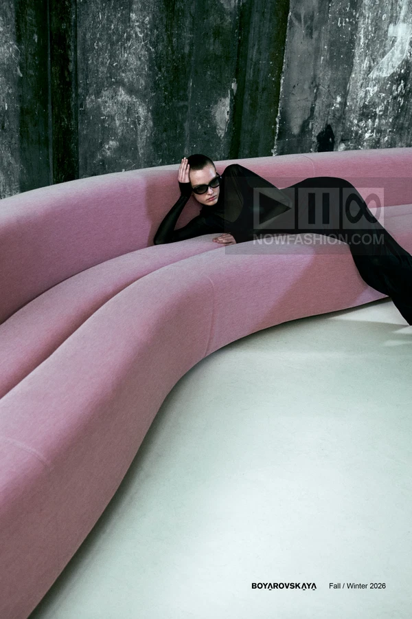 A person in a black long-sleeve top and black trousers reclines on a curved pink sofa against a textured gray wall.