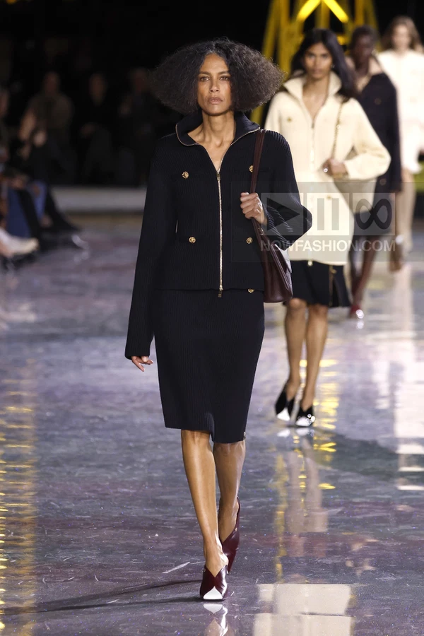 Model walking in a black ribbed knit jacket and matching midi skirt with a dark red shoulder bag and mules.