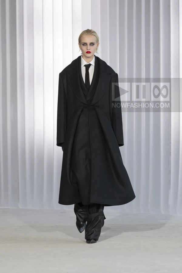 Model wearing a black oversized wool coat over a white shirt and black tie, with wide-leg black trousers on a runway.