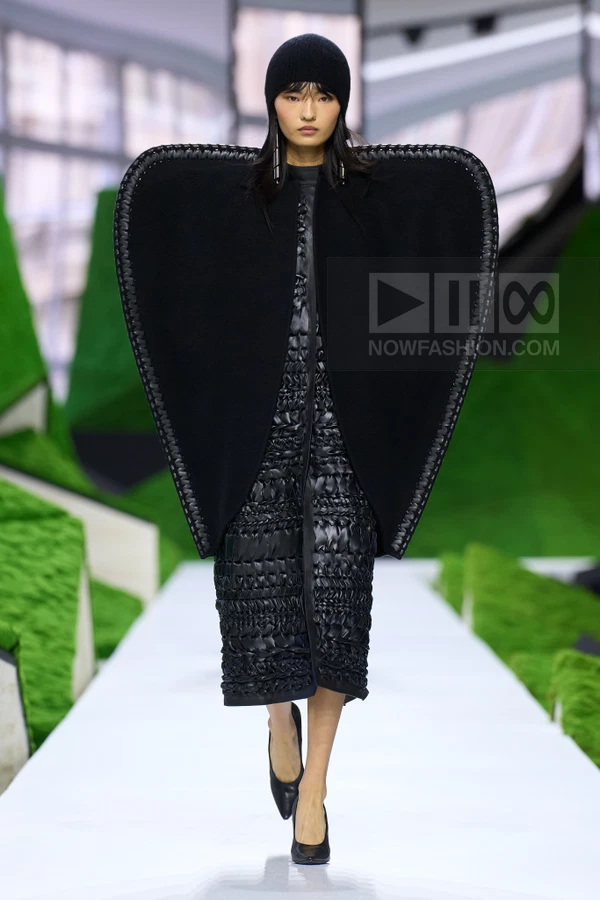 Model wearing a black oversized cape with leather trim over a quilted midi dress and a black beanie on a runway.