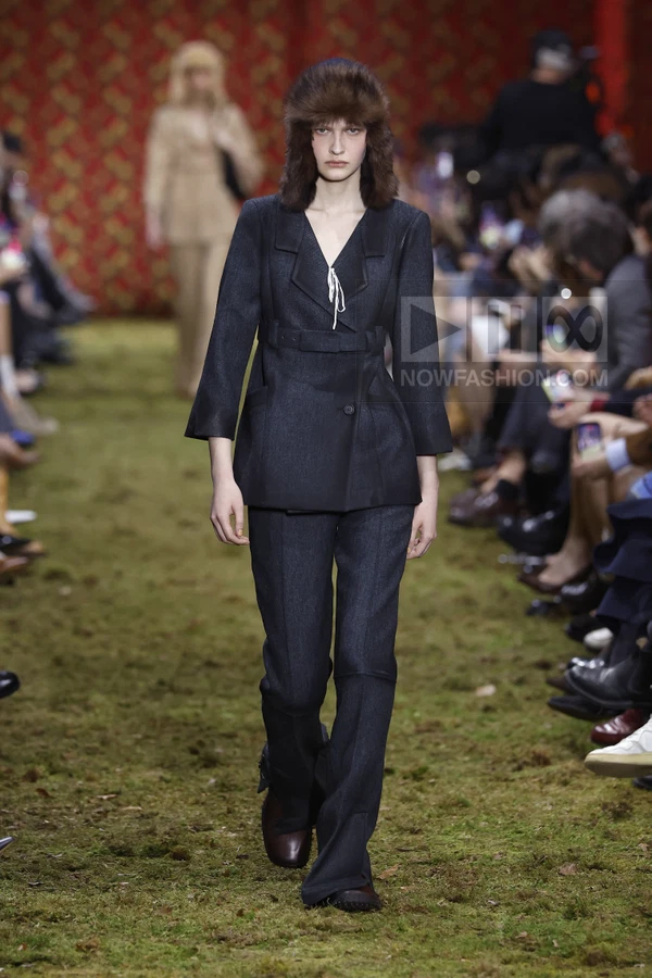 Model walking in a dark gray wool suit with a belted jacket and fur trapper hat on a mossy runway.